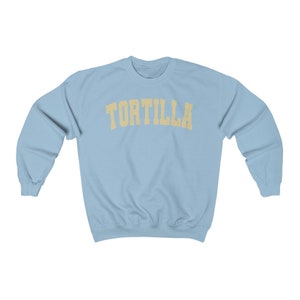 Puede incluir: Una sudadera azul claro con la palabra "TORTILLA" impresa en un arco amarillo sobre el pecho.