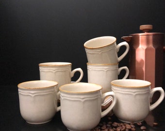 May include: Six white ceramic coffee mugs with gold trim, arranged in a semi-circle on a dark surface. A copper coffee pot is visible in the background.