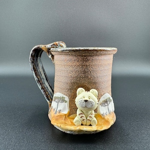May include: Handmade ceramic mug with a textured brown exterior and a decorative handle. The mug features a sculpted, light-colored tiger figure with brown stripes. Two small, textured rock-like elements flank the tiger.