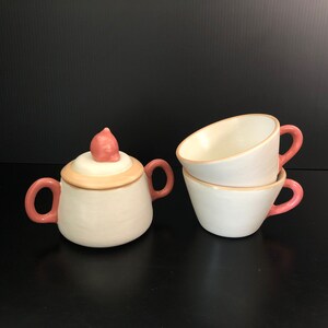 May include: A ceramic tea set featuring a sugar bowl with a lid and two teacups. The set is primarily white with pink handles and accents. The sugar bowl has a unique, rounded shape with a pink knob on the lid. The teacups are stacked and have pink handles.