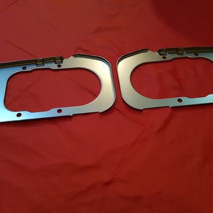 May include: Two silver metal automotive parts with rounded edges and multiple holes. The parts are on a red surface.