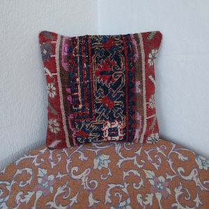 Puede incluir: Cojín decorativo cuadrado con diseño patchwork. El cojín presenta una sección central con un patrón azul oscuro y rojo, enmarcado por bordes en tonos rojos, marrones y rosas. El cojín está colocado sobre una superficie estampada.