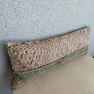 May include: A rectangular cushion with a vintage rug design. The cushion features a faded pink and beige central pattern, bordered by a green band. The cushion is placed on a light wooden surface.