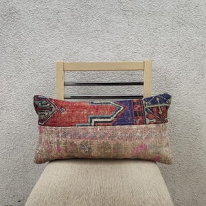 May include: A rectangular decorative pillow with a patchwork design. The top section features a red and blue geometric pattern, while the bottom section has a faded, neutral-toned floral design. The pillow is resting on a wooden chair.