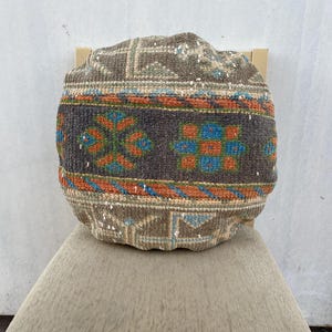 May include: A round, stuffed floor cushion featuring a textured, woven surface with a multi-colored geometric pattern. The design includes bands of gray, beige, orange, and blue, showcasing intricate, repeating motifs across the entire exterior.