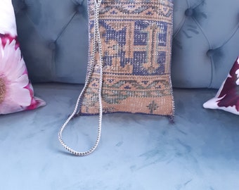 May include: A rectangular, woven bag with a geometric pattern in shades of blue, tan, and green. The bag has a long, silver-colored strap. The bag is sitting on a blue surface.