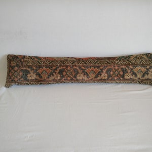 May include: A long, rectangular pillow with a detailed, patterned design. The pillow features a mix of colors, including dark green, rust, and beige, creating a vintage aesthetic. The pattern appears to be inspired by traditional rug designs.
