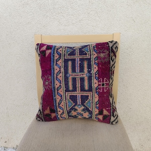 Può includere: Un cuscino quadrato decorativo con un motivo geometrico. Il cuscino presenta uno sfondo magenta intenso con un design blu navy centrale e accenti di verde acqua, pesca e nero. Il cuscino è posto su una sedia beige.