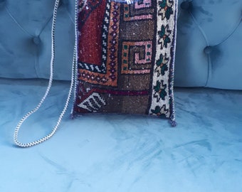 May include: A rectangular shoulder bag with a woven, patterned design in shades of burgundy, brown, and cream. The bag features a white rope strap and is displayed on a blue surface. The bag's pattern includes geometric shapes and floral motifs.