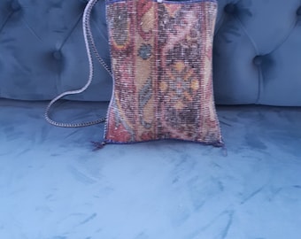 May include: A rectangular, patterned bag with a woven strap. The bag features a colorful, intricate design with shades of red, blue, and gold. The bag is resting on a blue surface.