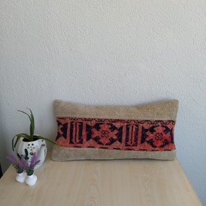 Può includere: Cuscino rettangolare beige con una fascia centrale di motivi geometrici rossi e blu navy. Il cuscino è posto su una superficie in legno chiaro, con un muro bianco sullo sfondo. Una piccola pianta in vaso e vasi decorativi sono a sinistra.