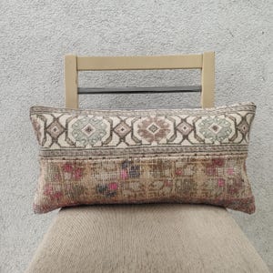 Op de afbeelding: Rechthoekig sierkussen met een vintage tapijtontwerp. Het bovenste gedeelte heeft een herhalend geometrisch patroon in beige, bruin en groen. Het onderste gedeelte heeft een bloemenpatroon met roze en bruine tinten. Het kussen ligt op een stoel.