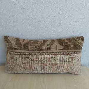 Op de afbeelding: Rechthoekig decoratief kussen met een vintage tapijtontwerp. Het kussen heeft een bruin en beige geometrisch patroon aan de bovenkant, een roze en beige bloemenrand en een beige basis. Het ligt op een licht houten oppervlak.