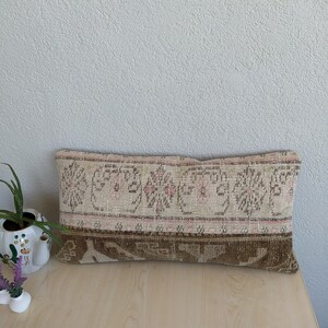 Op de afbeelding: Rechthoekig decoratief kussen met een beige en bruin patroon. Het kussen heeft een centrale band van beige met roze bloemmotieven en een onderste band van bruin met een geometrisch patroon. Het ligt op een houten oppervlak.