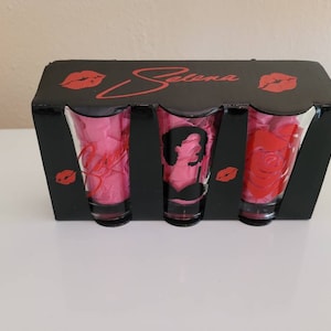 May include: A black box set containing three shot glasses. The glasses feature images of a silhouette, a rose, and the word "Bidi Bidi Bom Bom". The box has the name "Selena" in red script and red lip prints.