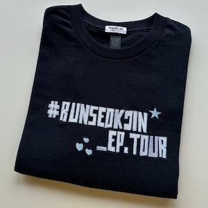Op de afbeelding: Zwart T-shirt met ronde hals en de tekst "#RUNSEOKJIN _EP.TOUR" in zilveren glitterletters. Een blauwe ster en drie blauwe hartjes staan ook op de shirt. Het shirt is netjes opgevouwen.