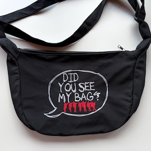 May include: A black crossbody bag featuring an embroidered white speech bubble with the text "DID YOU SEE MY BAG?" and a row of small red figures standing at the bottom. The bag has a top zipper closure and an adjustable black shoulder strap.