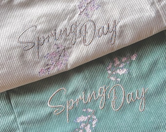 May include: Two fabric swatches with the text "Spring Day" embroidered in brown thread. The top swatch is cream colored and the bottom swatch is a light green.