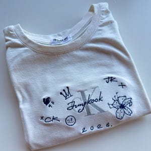 Könnte beinhalten: Ein cremefarbenes Kurzarm-T-Shirt mit dunkelblauen Stickereien, darunter eine Krone, eine Blume, ein zwinkerndes Gesicht, ein Herz mit der Zahl 97, der Text "Jungkook", "JK", "CK", "2026" sowie ein großer, silberfarben gestickter Buchstabe K.