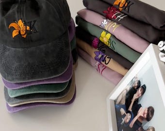 May include: A stack of six t-shirts with embroidered flowers and initials. The shirts are in various colours, including grey, pink, green, and beige. A black baseball cap with an embroidered flower and the initials "JK" is also pictured.