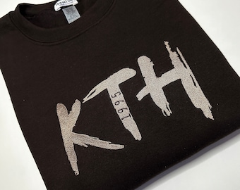 Puede incluir: Una sudadera marrón con un logotipo bordado en plata que dice "KTH 1996".