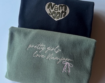 Puede incluir: Dos sudaderas dobladas, una azul marino y otra verde oliva. La sudadera azul marino tiene un corazón bordado con el nombre "Namjoon". La sudadera verde oliva tiene el texto bordado "pretty girls Love Namjoon" con un lazo rosa.