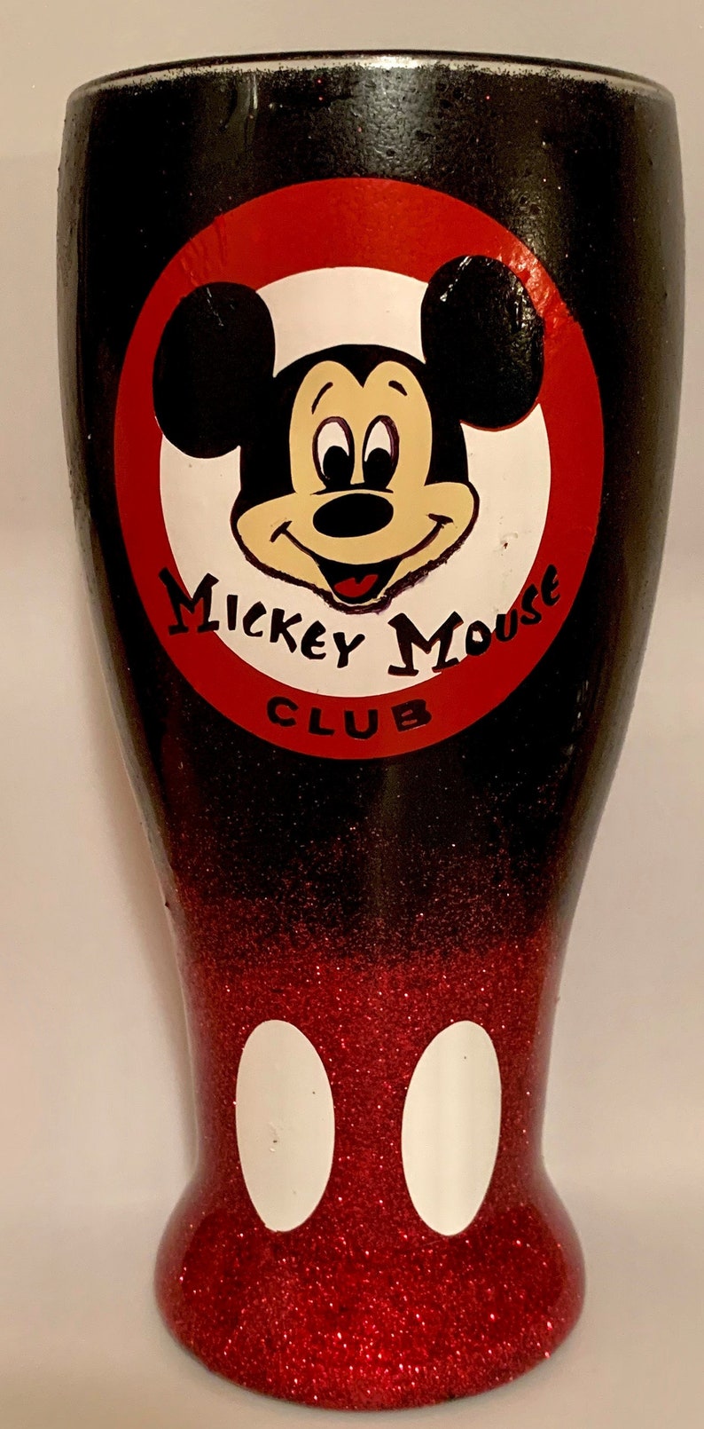 May include: A black and red glitter tumbler with a white Mickey Mouse design. The design includes Mickey Mouse's face with the words "Mickey Mouse Club" around it. The bottom of the tumbler has two white circles.