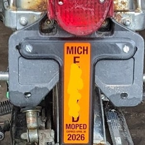 May include: A close-up of a black and orange Michigan moped license plate with the text "MICH" and "MOPED EXPIRES APRIL 30 2026".