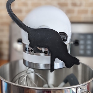 May include: A black cat silhouette kitchen utensil holder attached to a white KitchenAid stand mixer.