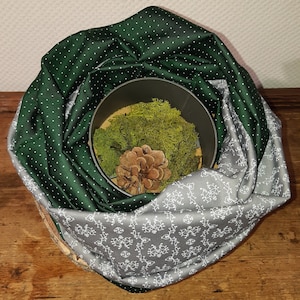 Könnte beinhalten: Dekorative Anordnung mit einem dunkelgrünen Stoff mit weißen Punkten und einem grauen Stoff mit weißem Blumenmuster. Im Inneren befindet sich ein Metallbehälter, gefüllt mit grünem Moos und einem Tannenzapfen.