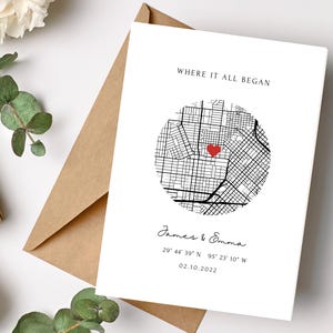 May include: A white card with a map design and the text "WHERE IT ALL BEGAN." The map features a red heart marking a location. Below the map, the names "James & Emma," coordinates, and the date 02.10.2022 are printed. The card is next to a brown envelope.