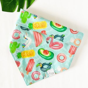 May include: A light blue bandana with a summer-themed print featuring inflatable pool floats, including a flamingo, a pineapple, an avocado, a unicorn, a watermelon, and a popsicle. The bandana has a white label with the text "Calico & Co."