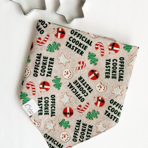May include: A beige bandana with a repeating pattern of Christmas cookies, candy canes, and snowmen. The text "Official Cookie Taster" is printed on the bandana.