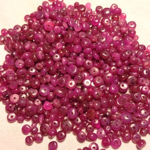 Puede incluir: Un montón de pequeñas cuentas de rubí redondas y pulidas de varios tamaños. Las cuentas son de un color rosa rojizo intenso y rico, con agujeros para ensartar. Las cuentas están esparcidas sobre una superficie blanca.