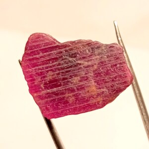 May include: A close-up of a pink ruby gemstone with striations. The ruby is held by tweezers against a white background.