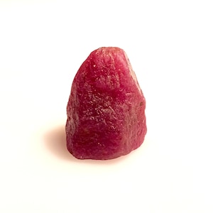 Peut inclure: Une pierre précieuse de rubis brute et non taillée. Le rubis est d'un rouge profond et riche avec une surface texturée. La pierre est posée sur un fond blanc, soulignant sa forme et sa couleur naturelles.