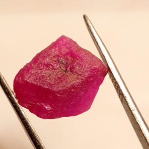 May include: A vibrant pink gemstone with a rough, natural finish. The gemstone is held in tweezers, showcasing its irregular shape and unique character.