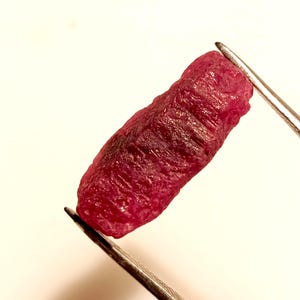 Puede incluir: Primer plano de una piedra preciosa de rubí rojo oscuro sostenida por unas pinzas. El rubí es una piedra en bruto sin pulir con una superficie estriada.