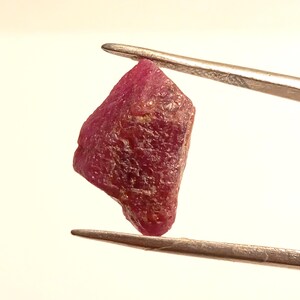 May include: A deep red ruby gemstone with a rough, faceted surface. The ruby is held by tweezers against a white background.