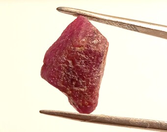 May include: A deep red ruby gemstone with a rough, faceted surface. The ruby is held by tweezers against a white background.