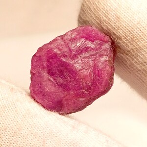 May include: A rough, uncut ruby gemstone with a deep pinkish-red hue. The natural stone has a textured surface and irregular shape, showcasing its raw, unpolished state. The ruby is set against a white background.