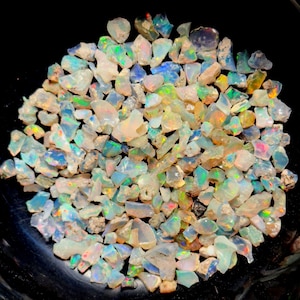 Puede incluir: Una colección de pequeñas virutas de ópalo iridiscente de varias formas y tamaños. Las virutas son una mezcla de blanco, azul, verde y naranja.
