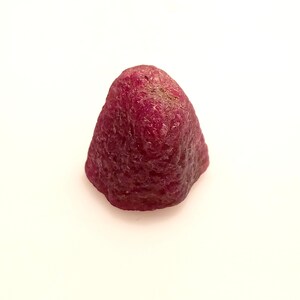 May include: A deep red ruby gemstone with a rough, natural surface. The ruby is a triangular shape with a pointed tip.