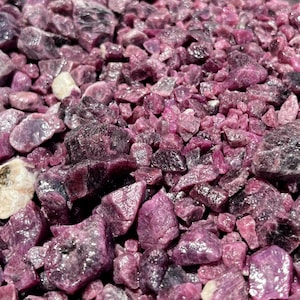 May include: A collection of raw, unpolished ruby gemstones in varying shades of deep magenta, purple, and pink. The stones have irregular, jagged shapes and faceted surfaces that reflect light, with a few small white and tan mineral inclusions.
