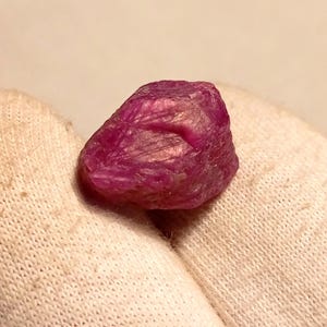 May include: A raw, unpolished ruby gemstone. The ruby is a deep, saturated pinkish-red colour with a textured surface. The gemstone is resting on a white fabric.