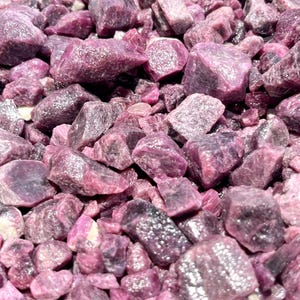 May include: A collection of raw, unpolished ruby gemstones in varying shades of deep magenta, purple, and dark pink. The stones feature irregular, jagged shapes with natural crystalline textures, some showing translucent facets and opaque surfaces.