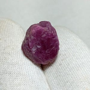 Könnte beinhalten: Ein rauer, ungeschliffener Rubin-Edelstein. Der Rubin hat eine tiefe, gesättigte Magenta-Farbe mit einer leicht unregelmäßigen, strukturierten Oberfläche. Der Edelstein wird vor einem weißen Hintergrund gezeigt.