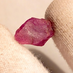 Puede incluir: Una gema de rubí en bruto, sin pulir, que muestra un color magenta intenso. La gema se sostiene entre dos superficies de tela blanca, resaltando su forma natural e irregular y su textura cristalina. La iluminación enfatiza las facetas y la estructura interna de la piedra.