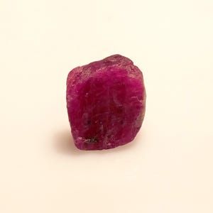 Puede incluir: Una piedra preciosa de rubí rosa oscuro con una forma natural y rugosa. El rubí es opaco y tiene una superficie ligeramente texturizada.