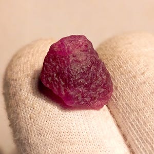 Peut inclure: Une pierre précieuse brute de rubis non taillée, de couleur magenta foncé. Le rubis est présenté sur un fond de tissu blanc, mettant en valeur sa forme naturelle irrégulière et sa surface texturée. Ce joyau est un minéral brut naturel.